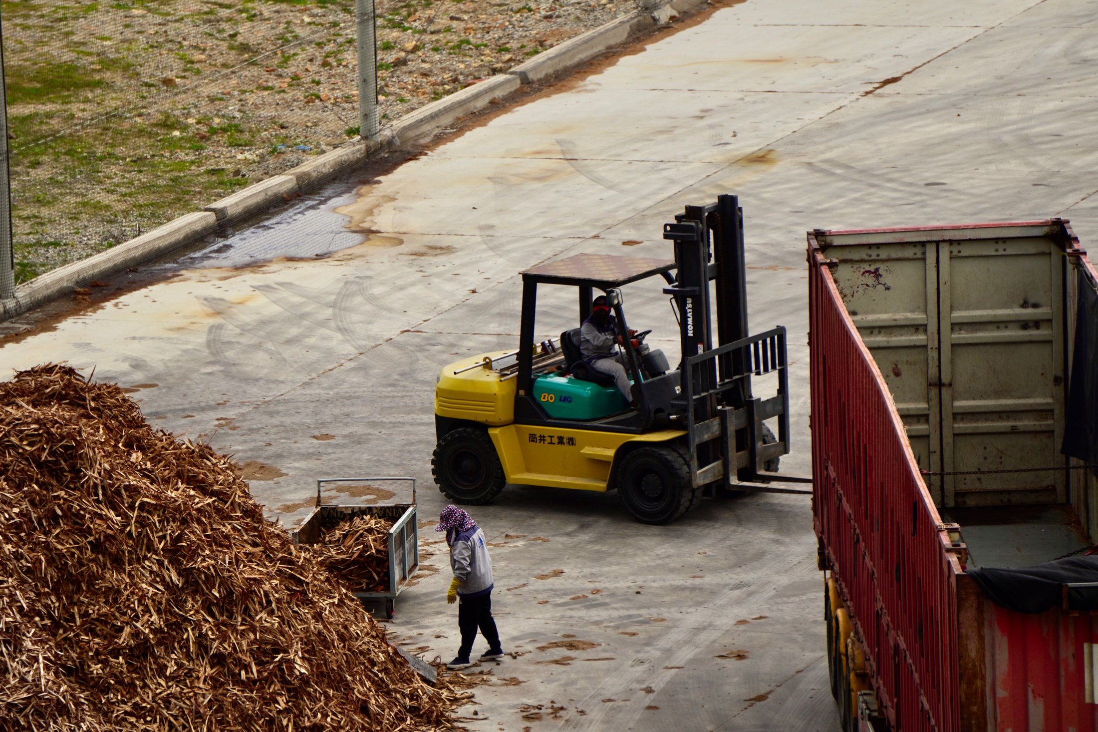 An operator drives a yellow and green forklift beside an open container and a pile of wood chips in an industrial yard.