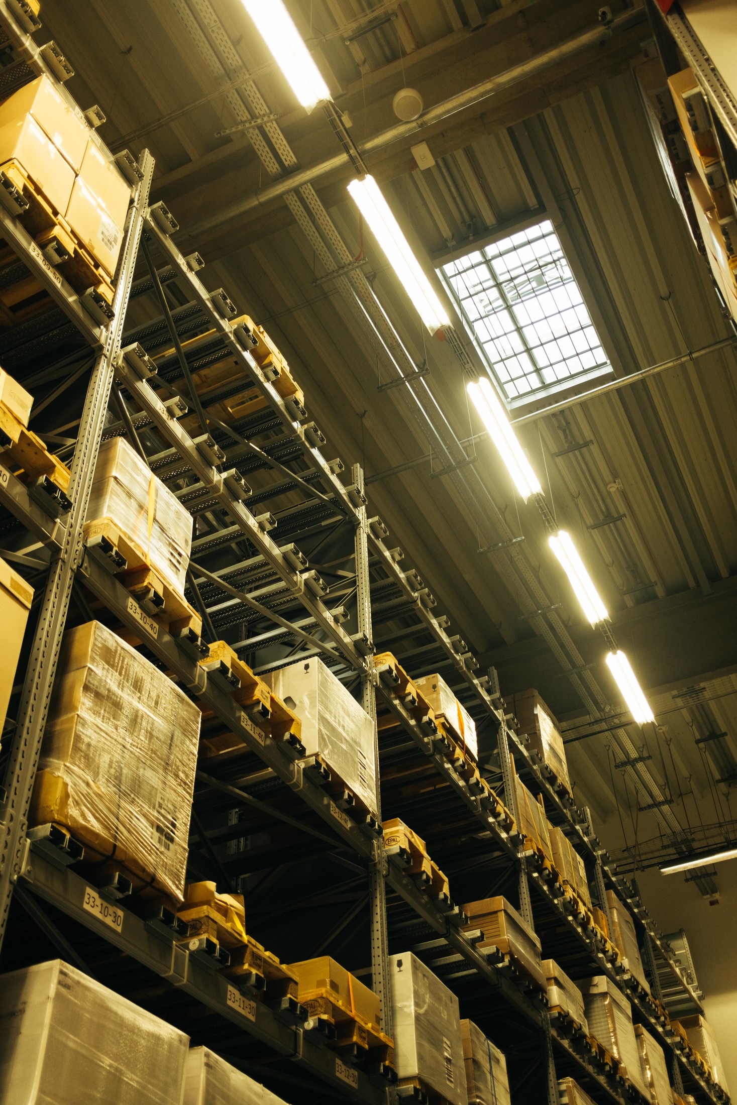 Tall warehouse racks hold wrapped pallets and cartons beneath overhead lights and a skylight.