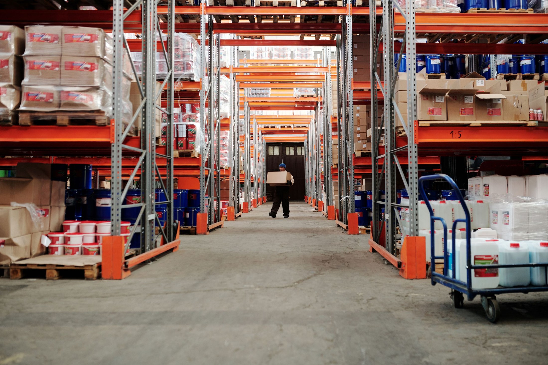 A worker carries a cardboard box down the center aisle between stocked warehouse racks.